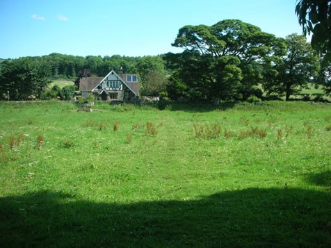 Photograph of a grass field with a house building in the background, there is no visible path or disturbance of the ground.