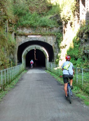 Two cyclists heading into a tunnel through a rock face on tarmac roads.